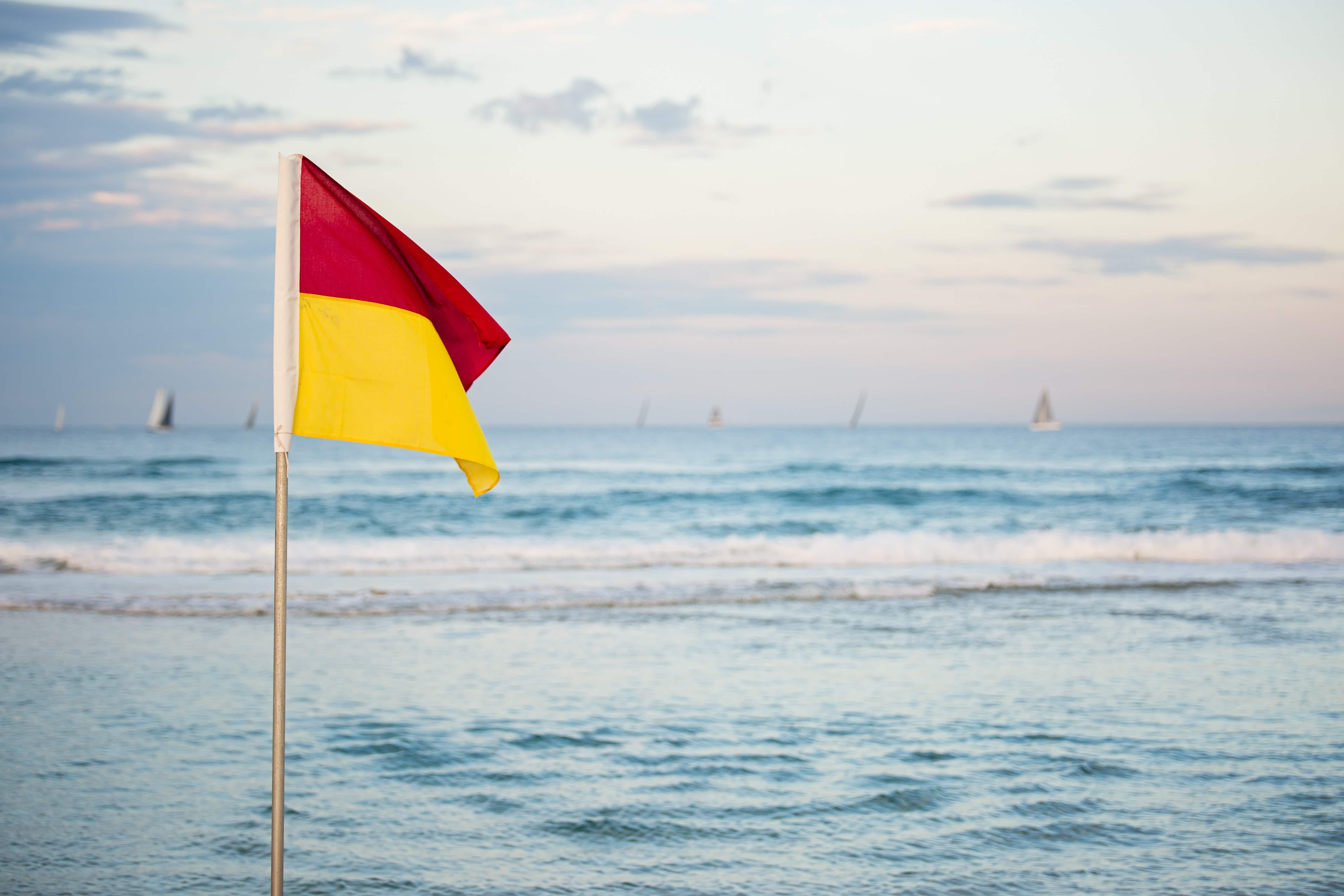 Red and yellow flag on a beach - yachts and sale boats in the background at dusk