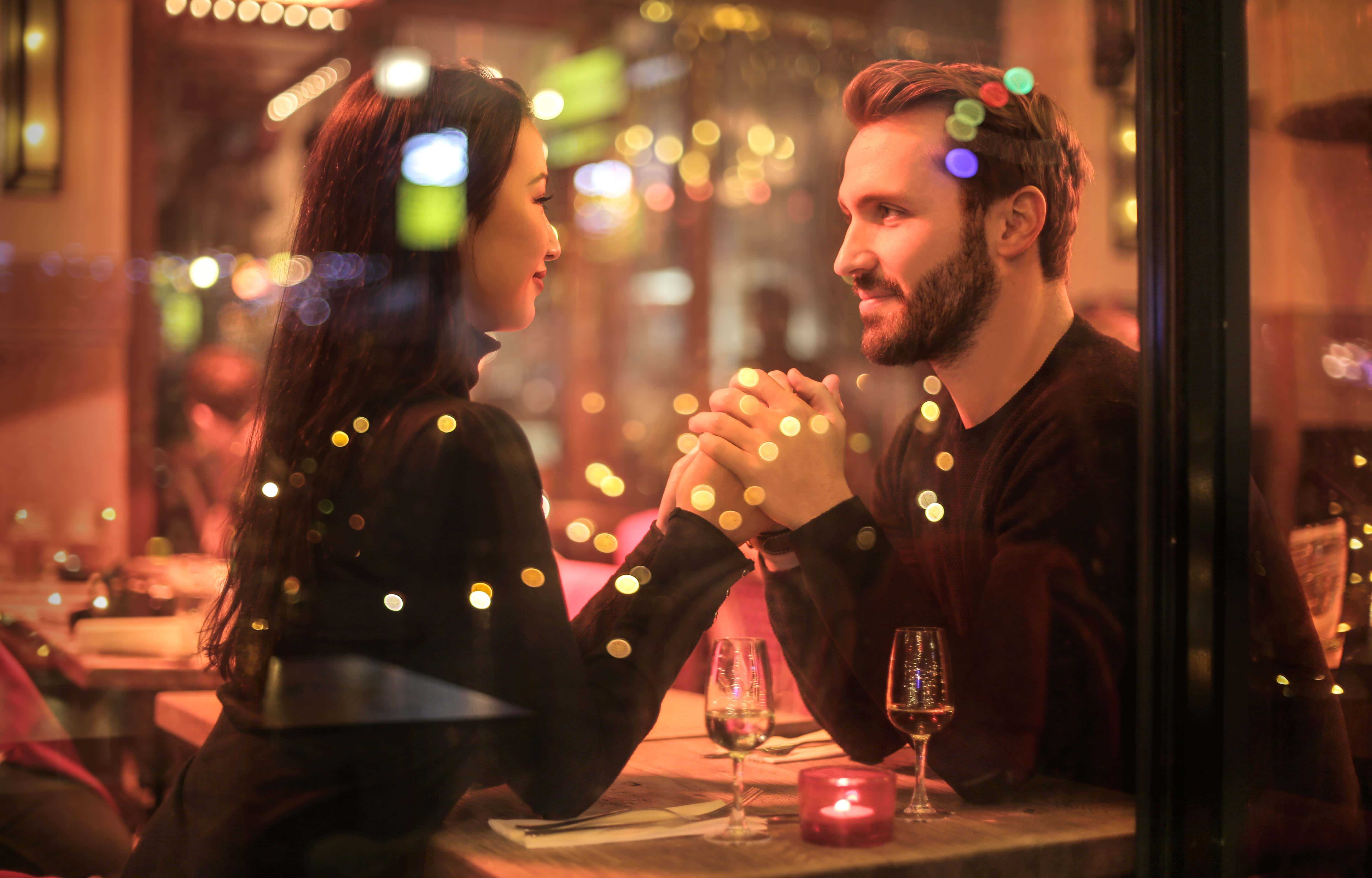 A young couple holds hands and looks into each others eyes over a candle lit dinner